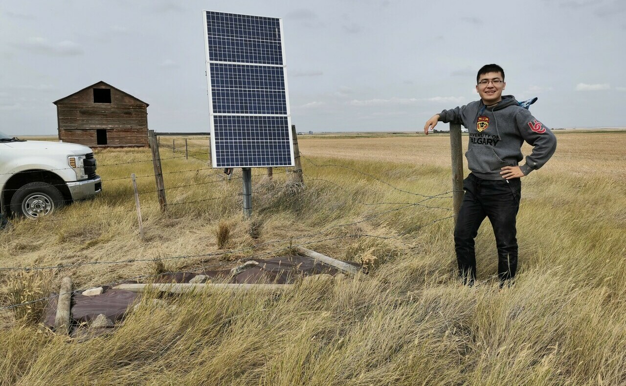 Jingchuan Wang (far right) poses in a grass field next to a solar cell, a white truck, and a wood structure