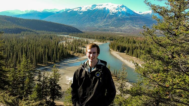 Evan stands in front of valley with river cutting through trees, a mountain range in distance behind